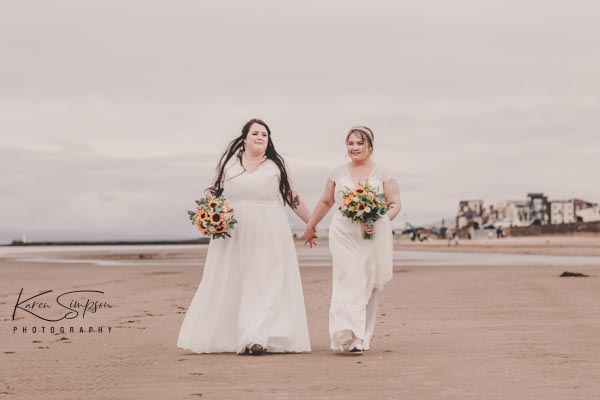 Two brides walking along Ayr Beach holding hands